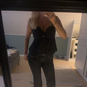 Svart ärmlös blus med knyt - Supersöt svart blus med broderade hål och knyt framtill. Toppen är ärmlös och har en lätt volangkant nedtill. Perfekt för dig som gillar en feminin och luftig stil. Snygg till både jeans och kjol!