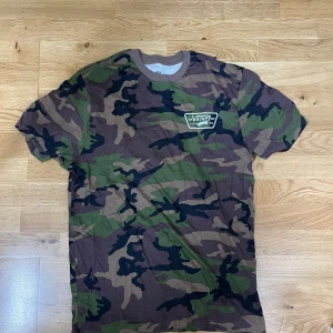 Camo t-shirt från Vans - Säljer en kortärmad t-shirt från Vans i klassiskt camouflage-mönster med gröna, bruna och svarta toner. T-shirten har rund halsringning med ribbad kant och en liten Vans-logga på bröstet. Perfekt för dig som gillar streetwear och vill sticka ut.