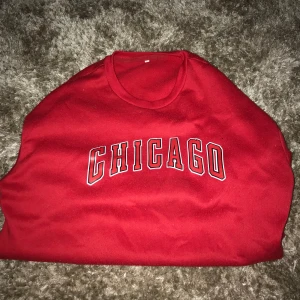 Röd sweatshirt med CHICAGO-tryck - Röd sweatshirt med coolt CHICAGO-tryck i vitt och svart på bröstet. Klassisk rund halsringning och avslappnad passform, perfekt för en sportig och trendig look. Materialet känns mjukt och bekvämt mot huden.