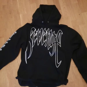 Svart Revenge hoodie med tryck - Svart hoodie från Revenge med stort vitt logotryck på bröstet och en stor vit hand på ryggen. Hoodien har huva, långa ärmar och känguruficka. Snygg streetwear-stil med tryck även på ena ärmen. Perfekt för dig som gillar bold statement-plagg.