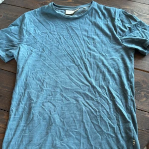 Blå t-shirt från Tailored Originals - Snygg blå t-shirt från Tailored Originals i klassisk modell. T-shirten är kortärmad och har en rund halsringning. Materialet känns mjukt och skönt, perfekt för en avslappnad stil. Diskret logga på bröstet ger en clean look.