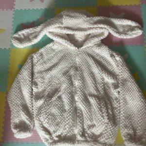 Vit teddyjacka med kaninöron - Supermysig vit teddyjacka med huva och långa kaninöron. Jackan har fluffig struktur, dragkedja framtill och är perfekt för dig som vill ha något gulligt och bekvämt.