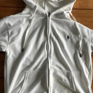 Vit Ralph lauren tröja - Vit tröja från Polo Ralph Lauren med dragkedja framtill, två fickor och klassisk logga på bröstet. Jackan har huva med snörning och är tillverkad i mjuk bomull. Perfekt för en chill och stilren look. Har inget kvitto 