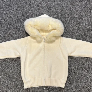 Vit Alessa fur jacket  - Mysig beige stickad jacka med huva och fluffig päls runt kanten. Jackan har dragkedja framtill och breda ribbade muddar vid ärmslut och nederkant. Perfekt för kalla dagar när du vill vara både varm och snygg.