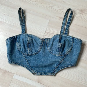Jeans korsettopp från NA-KD - Cool jeans korsettopp från NA-KD i klassisk blå denim. Toppen har markerade kupor, breda axelband och dragkedja bak. Snygga sömdetaljer och croppad passform ger en edgy vibe. Perfekt att styla med höga byxor eller kjol för en trendig look.