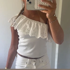 Vit one shoulder topp med volang - Superfin vit topp med one shoulder-design och bred volang med broderade blommor. Toppen är ärmlös och har en croppad passform, perfekt för varma dagar.
