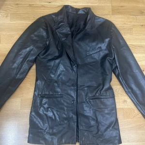 Svart skinnkavaj med knappar - Snygg svart kavaj i skinn med klassisk krage och tre knappar framtill. Jackan har två fickor nertill och en bröstficka, perfekt för en stilren look. Långärmad modell med slät finish och rak passform.