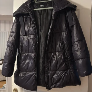Svart pufferjacka från ONLY - Säljer en svart pufferjacka från ONLY med huva och dragkedjor på fickorna. Jackan är quiltad och har en glansig finish, perfekt för kalla dagar. Den har lång ärm och en normal passform. Snygg och enkel att matcha med allt.