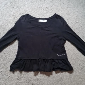 Svart volangtröja från Bondelid - Svart långärmad tröja från Bondelid med volangkant nedtill och broderad logga i vitt. Tröjan har rund halsringning och är gjord i mjuk bomull, perfekt för dig som gillar en enkel men ändå söt stil. Kan gå ned i pris vid snabb affär!