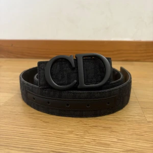 Svart  med CD-spänne - Snyggt svart bältemed stort mattsvart CD-spänne. Bältet är gjort i skinn. Perfekt accessoar för att lyfta din outfit.
