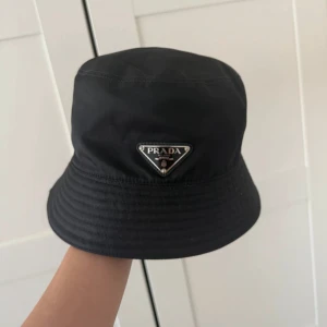 Svart Prada hatt  - Stilren svart bucket hat från Prada med klassisk triangellogo framtill. Hatten är köpt i Prada butiken i Strøget har haft den i ett år gillar ej den mer den är fotfarandre i bra skick hojta till för förhandlingar 
