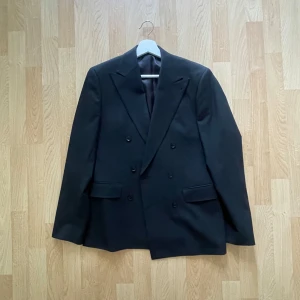 Svart kavaj från Bläck, tailored fit - Dubbelknäppt svart kavaj från Bläck med skräddad passform. Klassisk design med breda slag, två fickor med lock och innerfoder. Perfekt för dig som gillar stilren och tidlös look.
