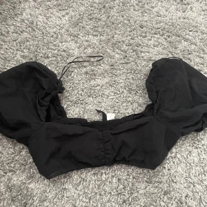 Svart croppad topp från H&M Divided - Supersnygg svart croppad topp från H&M Divided med puffiga korta ärmar och smockad rygg. Toppen har en söt knytning upptill och är perfekt för dig som gillar trendiga detaljer och en feminin look. Tillverkad i mjuk bomull som känns skön mot huden.