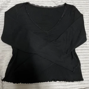 Svart långärmad v-ringad topp - Svart långärmad topp med v-ringning och spetskant vid halsen. Ärmsluten och nederkanten har en vågig, lätt ruffad finish. Tunt, stretchigt material som känns mjukt mot huden. Perfekt för dig som gillar en enkel men ändå snygg look.