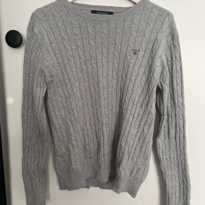 Grå kabelstickad tröja från GANT - Kabelstickad grå tröja från GANT i stretchig bomull. Klassisk rund halsringning, diskret broderad logga på bröstet och ribbade muddar. Perfekt för lager-på-lager och en clean look.