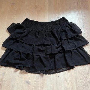Svart volangkjol - Supersöt svart kortkjol med flera volanger och bred resår i midjan. Kjolen har en lätt och luftig känsla, perfekt för att skapa en trendig och lekfull look. Passar till sneakers eller boots för en cool stil.