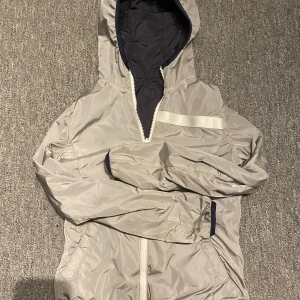 Colmar vindjacka i grått och blått - Snygg och lätt vindjacka från Colmar med huva och dragkedja. Perfekt för blåsiga dagar när du vill ha en stilren look. Jackan går att vända ut och in, så man kan ha den i grått eller i blått.