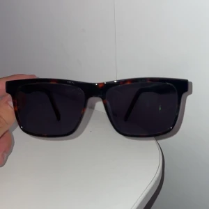 Svarta solglasögon från Hugo Boss, med 1.5 Styrka - Snygga solglasögon från Hugo med svart och röd båge samt mörka rektangulära glas. Bågarna har diskreta bruna detaljer och loggan i rött på sidan. Perfekt accessoar för att lyfta din stil i sommar.