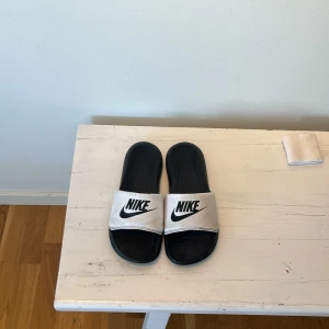 Svarta och vita Nike sandaler - Svarta sandaler från Nike med bred vit rem över foten och stor svart Nike-logga. Sval och enkel design med räfflad sula för extra grepp. Perfekta för sommardagar eller att chilla i efter träningen.