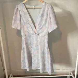 Pastellfärgad kortklänning från NLY Trend - Supersöt kortklänning från NLY Trend i pastelliga toner av ljusblått, rosa och vitt. Klänningen har v-ringning, puffiga korta ärmar och ett mjukt, abstrakt mönster. Perfekt för sommarens alla tillfällen och har en flowy passform.