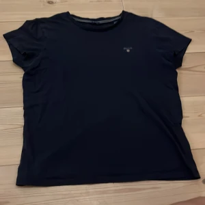 Svart t-shirt från GANT med logga - En stilren marin blå t-shirt från GANT med klassisk rund hals och diskret broderad logga på bröstet. T-shirten är i mjuk bomull och har en normal passform. Perfekt för dig som gillar enkel och tidlös stil. Och ett litet hål i vid ärmen