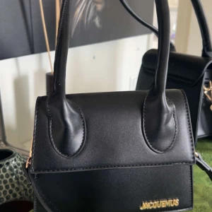 Svart Jacquemus handväska i skinn - Säljer en svart handväska från Jacquemus med stilren design och guldigt logotypdetalj framtill. Väskan har två rejäla handtag och är tillverkad i slätt skinn. Perfekt för dig som gillar minimalistisk och trendig look.