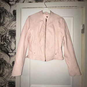 Ljusrosa skinnjacka med dragkedja - Snygg ljusrosa skinnjacka med guldiga detaljer och dragkedja framtill. Jackan har en kortare passform, lång ärm och spännen i midjan för extra stil. Perfekt för dig som vill ha en feminin touch på din outfit.