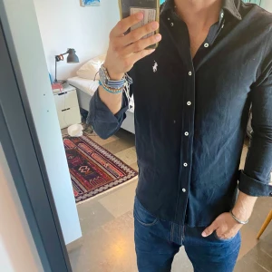 Svart skjorta från Ralph Lauren M - Säljer en svart skjorta från Ralph Lauren i storlek M. Skjortan har klassisk krage, vita knappar och den ikoniska broderade loggan på bröstet. Tillverkad i lätt bomullsmaterial som känns luftig och bekväm. Perfekt för dig som gillar stilren och tidlös look.