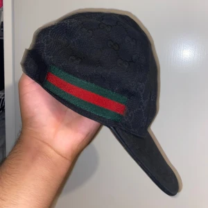Äkta Svart Gucci keps  - Snygg svart keps från Gucci med klassiskt GG-monogram över hela kepsen. På sidan finns den ikoniska gröna och röda randen. Justerbar rem baktill och böjd skärm. Perfekt för dig som vill ha en exklusiv streetstyle-look.