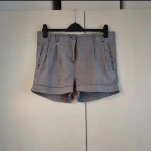 Shorts kort modell grå strl 38 - Ljusgråa shorts i använt skick men dom är fortfarande fina.  Shortsen har två fickor fram och två fickor bak, midjan är normal. Sydda veck framtill på shortsen. Benslut har en sydd kant. Shortsen har hällor för skärp, shortsen stängs med en knapp och gylf. INFO: Hund i hemmet. 