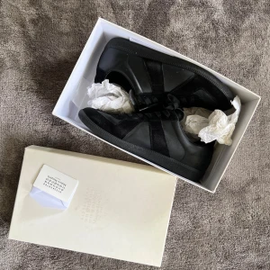 Svarta Maison Margiela sneakers i skinn och mocka - Maison Margiela sneakers i helsvart med en mix av skinn och mocka. Klassisk låg modell med rund tå, platt sula och svarta snören. Diskret logga på plösen och stilren design som passar till allt. Kommer med originalkartong och taggar.