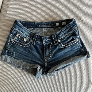 Blå jeansshorts från Miss Me, strl 27 - Säljer för att det är lite stora för mig har använt säker 2-3 gånger. Snygga blå jeansshorts från Miss Me med coola detaljer som grova vita sömmar, dekorativa knappar och broderad M på bakfickan. Shortsen har låg midja, uppvikta ben och slitningar för en edgy look. Perfekt för dig som gillar statement-plagg och denim. 