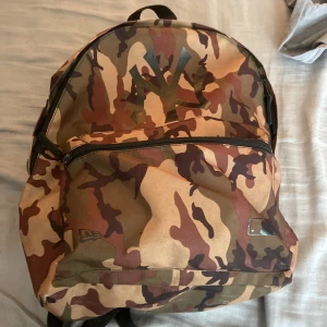 Camo ryggsäck New Era  - Säljer en fet ryggsäck från New Era med New York Yankees-logga. Väskan har camouflagemönster i brunt, beige och grönt, stor ytterficka med dragkedja och justerbara axelremmar. Perfekt för skolan eller stan, och rymmer allt du behöver. Kostar orginalt 500-400
