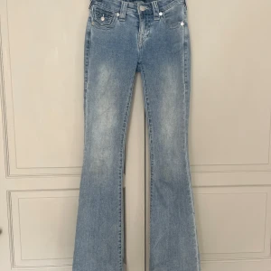 True Religion Becca Bootcut Jeans - Ljusblå bootcut jeans från True Religion, modell Becca Mid Rise Bootcut. Klassisk femficksdesign med snygga detaljerade bakfickor och silverfärgade knappar. Jeansen är i mjuk denim och har en lätt tvättad look för en avslappnad vibe.