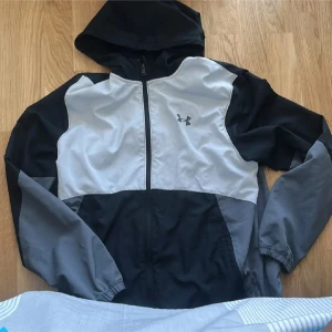 Under Armour kofta - Snygg vindjacka från Under Armour i svart, grått och ljusgrått. Jackan har huva, hel dragkedja och sportig design med logga på bröstet. Perfekt för utomhusaktiviteter och träning. Långärmad med elastiska muddar och lätt material som andas.