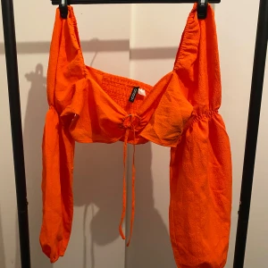 Orange croppad blus från H&M Divided - Säljer en knallorange croppad blus från H&M Divided. Toppen har långa puffärmar, smockad rygg och knyt framtill. Tunt, lätt material som känns luftigt och perfekt för varmare dagar. Supersnygg till höga jeans eller kjol!