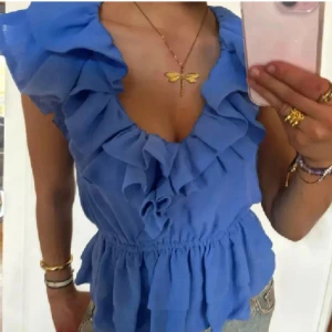Blå volangblus med v-ringning - Jättesöt blå blus med volanger längs v-ringningen och axlarna. Modellen är ärmlös och har en smockad midja som ger en snygg siluett.🩵