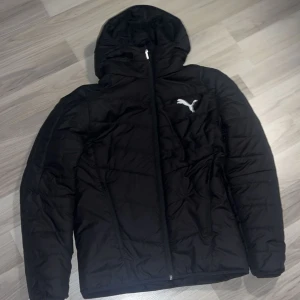 Svart pufferjacka från Puma - Svart pufferjacka från Puma med huva och dragkedja framtill. Jackan har ett stilrent vitt Puma-logo på bröstet och ryggen. Perfekt för kalla dagar, med quiltad design och två sidofickor. Snygg och enkel att matcha till det mesta.