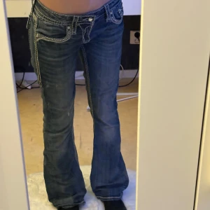 Rock Revival bootcut - Säljer ett par blå bootcut jeans från Rock Revival med låg midja. Jeansen har tydliga kontrastsömmar, snygga broderier och detaljer på fickorna. Materialet är klassisk denim och passformen är bootcut. Midja: 39cm rakt över. Innerben: 80cm och benöppning 23cm rakt över. ❤️skriv gärna vid frågor!