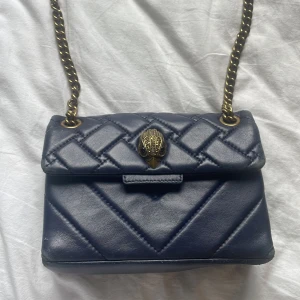 Mörkblå quiltad axelväska med kedja - Snygg mörkblå quiltad axelväska i skinn med guldfärgad kedjerem och unik uggleformad låsdetalj framtill. Väskan har en klassisk boxig form och coola sömmar i rutmönster. Perfekt för dig som gillar statement-detaljer och stilren design. ❤️