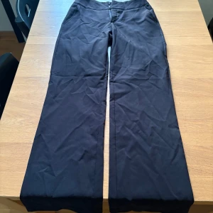 Svarta kostymbyxor från Madlady - Snygga svarta kostymbyxor från Madlady i storlek EU38. Byxorna har raka ben, mellan midja och stängs med dragkedja och två hyskor fram. Baktill finns två dekorativa fickor. Materialet är mjukt och har en lätt stretch för skön passform.