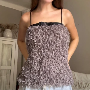 Ljuslila fluffig tubtopp med rosett - Unik tubtopp i ljuslila med fluffigt pälsliknande material och smala svarta axelband. Toppen har en svart rosett framtill som ger en extra söt touch. Perfekt för dig som vill sticka ut med en cool och trendig look.