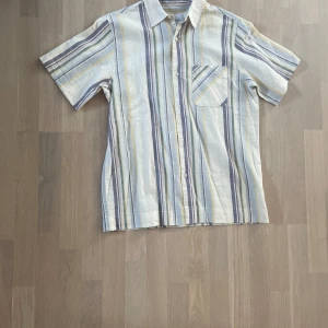 Randig kortärmad skjorta från Vailent - Snygg kortärmad skjorta från Vailent i ljusa färger med ränder i blått, gult och grått. Skjortan har klassisk krage, bröstficka och knäppning framtill. Perfekt för en chill sommardag när du vill vara både bekväm och stilren.