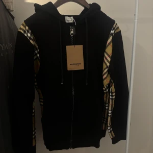 Burberry Hoodie med dragkedja – Nytt skick - Svart Burberry-hoodie med dragkedja och klassiskt rutmönster på ärmarna. Har bara legat i skåpet, aldrig använd. Nytt skick med tag kvar. Inga skambud, endast seriösa köpare.