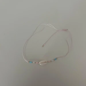 Sommrigt snäckhalsband med pärlor - Ett tunt halsband med ljusrosa snöre, dekorerat med små pärlor i blått, vitt och guld samt en snäcka i mitten. Perfekt accessoar för en strandig look och sommarkänsla.