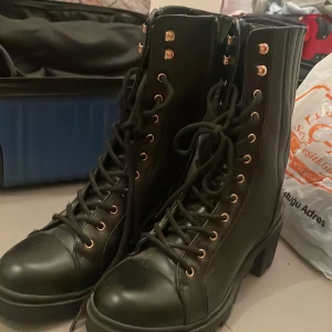 Svarta boots med snörning och klack - Snygga svarta boots med chunky klack och snörning hela vägen upp. Skorna har guldfärgade öljetter och är gjorda i ett slätt skinnliknande material. Perfekta för dig som vill ha en edgy och trendig look till hösten eller vintern.