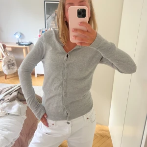 Ljusgrå kofta med knappar - En stilren ljusgrå kofta med rund hals och knäppning framtill. Koftan har långa ärmar och är tillverkad i ett mjukt, tunt material som känns skönt mot huden. Perfekt att slänga på sig över en t-shirt eller linne för en clean look.