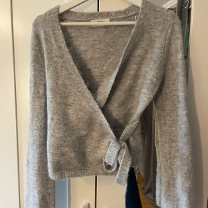 Grå omlottkofta från Gina Tricot - Supermjuk grå omlottkofta från Gina Tricot i ullblandning. Koftan har långa ärmar, v-ringning och knyt i sidan för en snygg och avslappnad look. Oanvänd, och slutsåld eftertraktad kofta från Gina!!