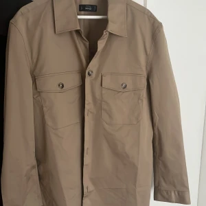 Beige overshirt från MNG Essentials - Snygg beige overshirt från MNG Essentials med klassisk krage och två stora bröstfickor med knappar. Jackan har raka linjer, lång ärm och stilren design som passar till många outfits. Perfekt för lager-på-lager och enkel att matcha. Nypris 500 säljer för 150