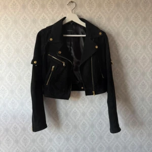 Svart mocka skinnjacka Rock'n Blue - Snygg svart jacka i mocka från Rock'n Blue med guldiga detaljer och dragkedjor. Croppad modell med bred krage och flera fickor framtill. Jackan har lång ärm. Perfekt för dig som vill ha en statement piece i garderoben. Knappt använd. Rökfritt hem.